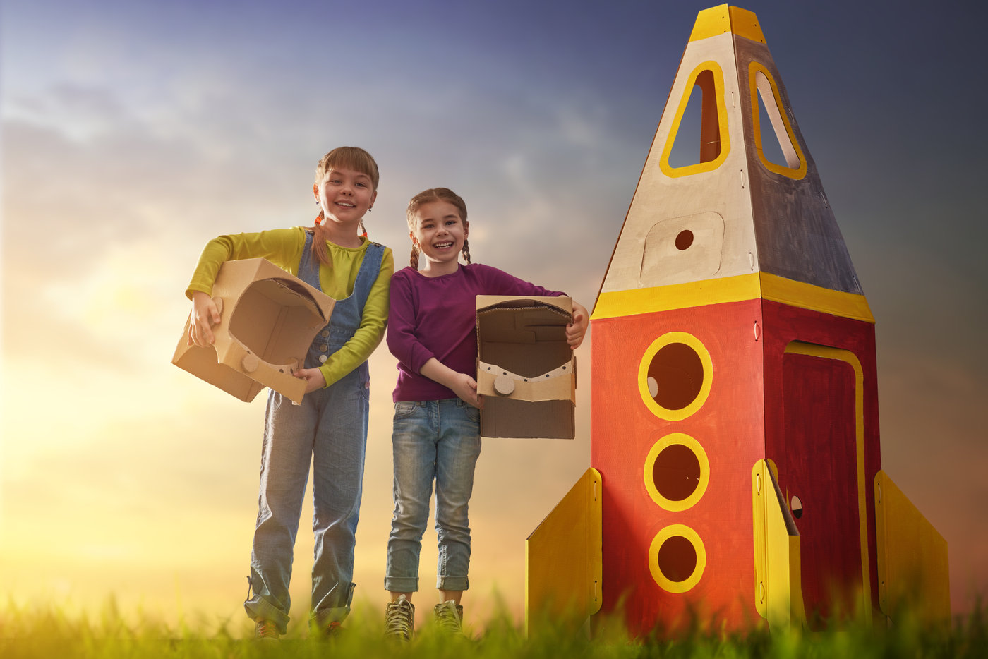 Zwei Kinder mit Kartonflügeln stehen neben einer selbstgebauten Rakete